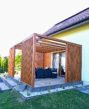 Drewniana konstrukcja z pionowych desek tworząca zadaszone patio z meblami ogrodowymi, otoczone zielenią i białym kamieniem.