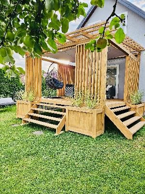Drewniana konstrukcja z pionowych listew, schodami i wiszącym fotelem na tle zielonego trawnika i domu.