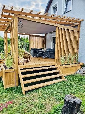 Drewniana konstrukcja zadaszonej werandy z meblami ogrodowymi, schodami i roślinami w donicach, przylegająca do jasnoszarego domu.