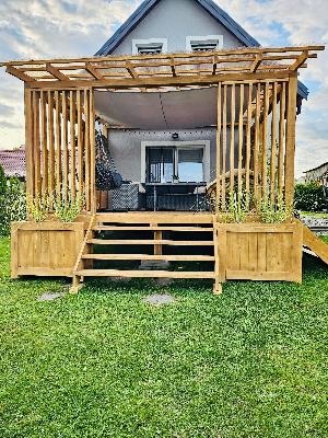 Drewniana altana z ażurowymi ścianami i schodami, donice z roślinami po bokach schodów, widok na dom w tle.