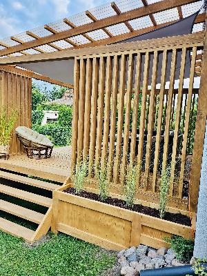 Drewniana konstrukcja z zadaszeniem i pionowymi panelami, osłaniająca taras z krzesłem w Górowo Iławeckie, z roślinami w donicy u podstawy.