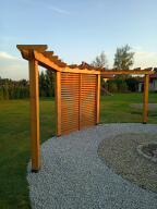Drewniana konstrukcja z przesłoną z lameli, tworząca wejście do ogrodu, wyłożonego jasnym żwirem, w tle zielony trawnik i drzewa pod błękitnym niebem.