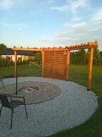 Drewniana konstrukcja altany z ażurową ścianą, otaczająca okrągłe palenisko wyłożone białym żwirem, na tle zielonego trawnika i zabudowań.