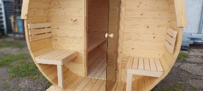 Drewniana sauna beczka z otwartymi drzwiami, widoczne wnętrze z ławkami, jasne drewno, naturalne oświetlenie.