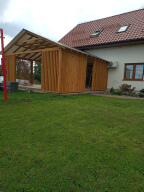 Drewniana konstrukcja przylegająca do budynku mieszkalnego z widocznym zadaszeniem i pionowymi deskami tworzącymi częściowe ściany, zielony trawnik na pierwszym planie, czerwony słup na skraju kadru.