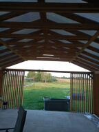 Zadaszona konstrukcja z drewnianym szkieletem i przezroczystym dachem, widok na pole i drzewa w oddali, pionowe drewniane listwy po bokach.