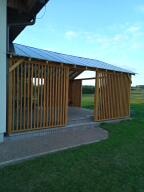 Drewniana konstrukcja z pionowych listew, prawdopodobnie zadaszenie tarasu lub wiata, z metalowym dachem, widok z boku na tle zielonego trawnika i błękitnego nieba.