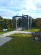 Nowoczesna, minimalistyczna konstrukcja z ciemnego drewna lub kompozytu, prawdopodobnie zadaszenie tarasu lub element dekoracyjny ogrodu, na tle zielonego trawnika i drzew. Widoczne ścieżki...