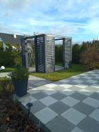 Nowoczesna altana z szarych, pionowych paneli na trawniku, z widocznym fragmentem szarej kostki brukowej w romby i błękitnym niebem w tle.