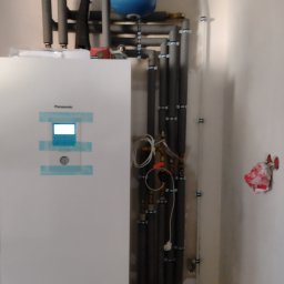 Łukasz Brewiński - Biała jednostka wewnętrzna z wyświetlaczem marki Panasonic zamontowana na ścianie, widoczne rury instalacji centralnego ogrzewania z izolacją termiczną i niebieski zbiornik wyrównawczy.