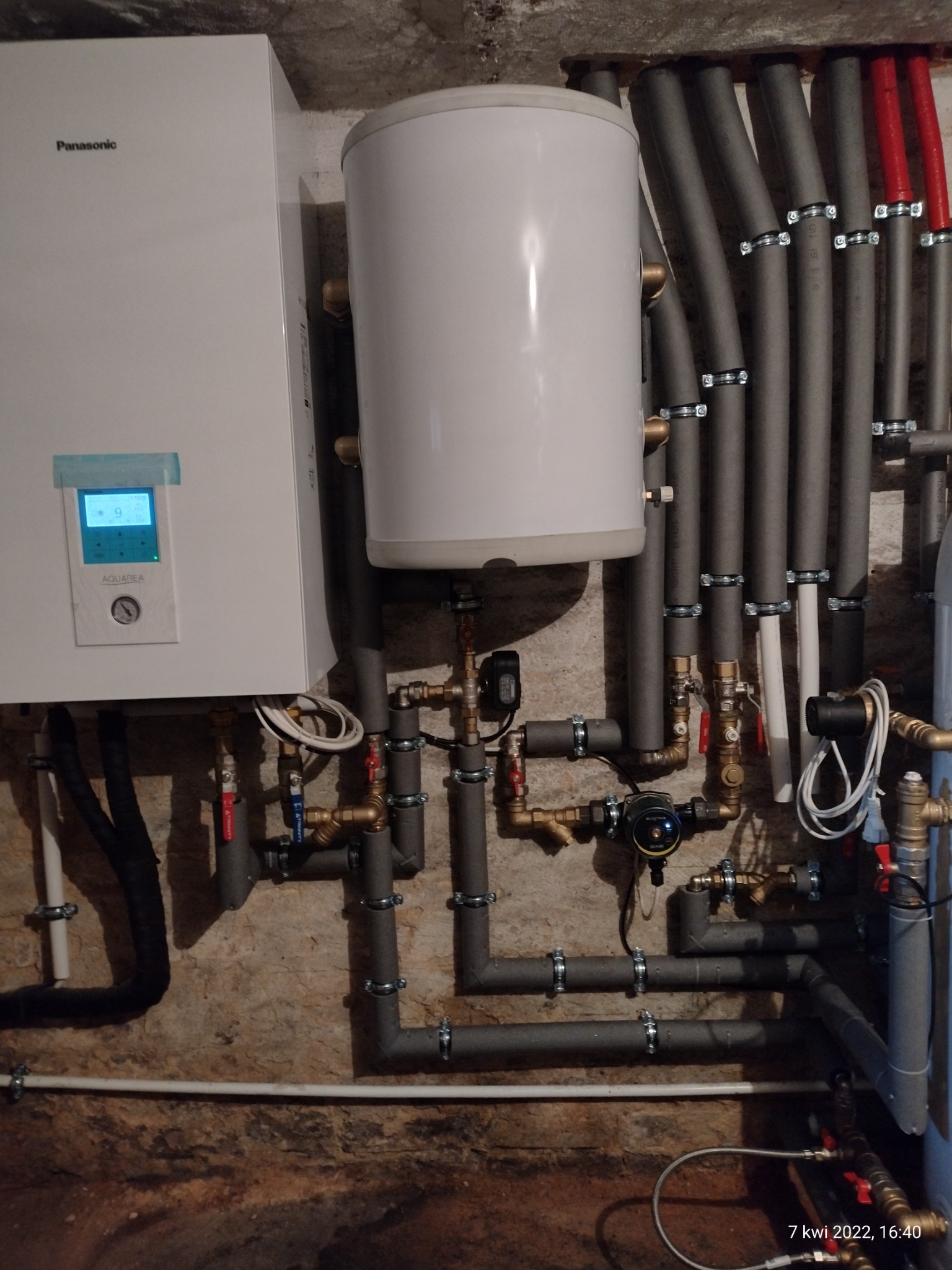 Biały zasobnik ciepłej wody użytkowej i panel sterowania Panasonic Aquarea zamontowane na ścianie z widocznymi rurami instalacji grzewczej w szarych izolacjach.