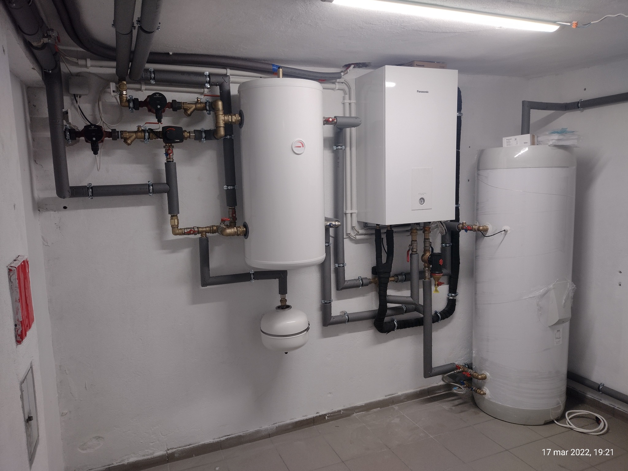 Białe zbiorniki i szare rury instalacji grzewczej w piwnicy, widoczne zawory, pompy obiegowe, naczynie przeponowe, całość oświetlona jarzeniówką.