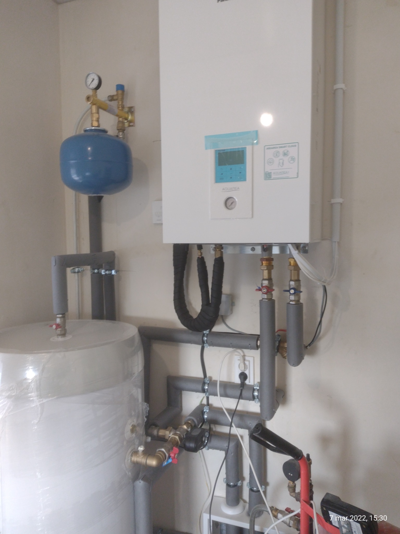 Biały bojler elektryczny z niebieskim zbiornikiem wyrównawczym i szarymi rurami instalacji grzewczej, widoczne złącza i manometr, instalacja w pomieszczeniu z jasnymi ścianami.