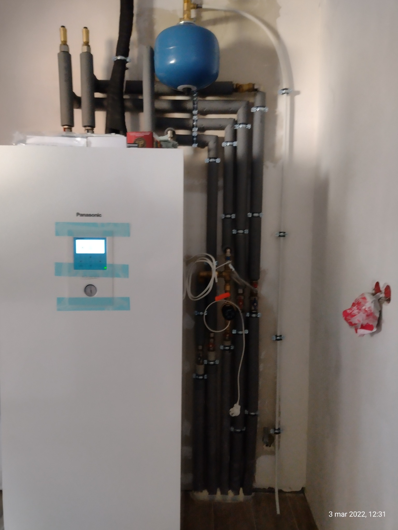 Biała jednostka wewnętrzna z wyświetlaczem marki Panasonic zamontowana na ścianie, widoczne rury instalacji centralnego ogrzewania z izolacją termiczną i niebieski zbiornik wyrównawczy.