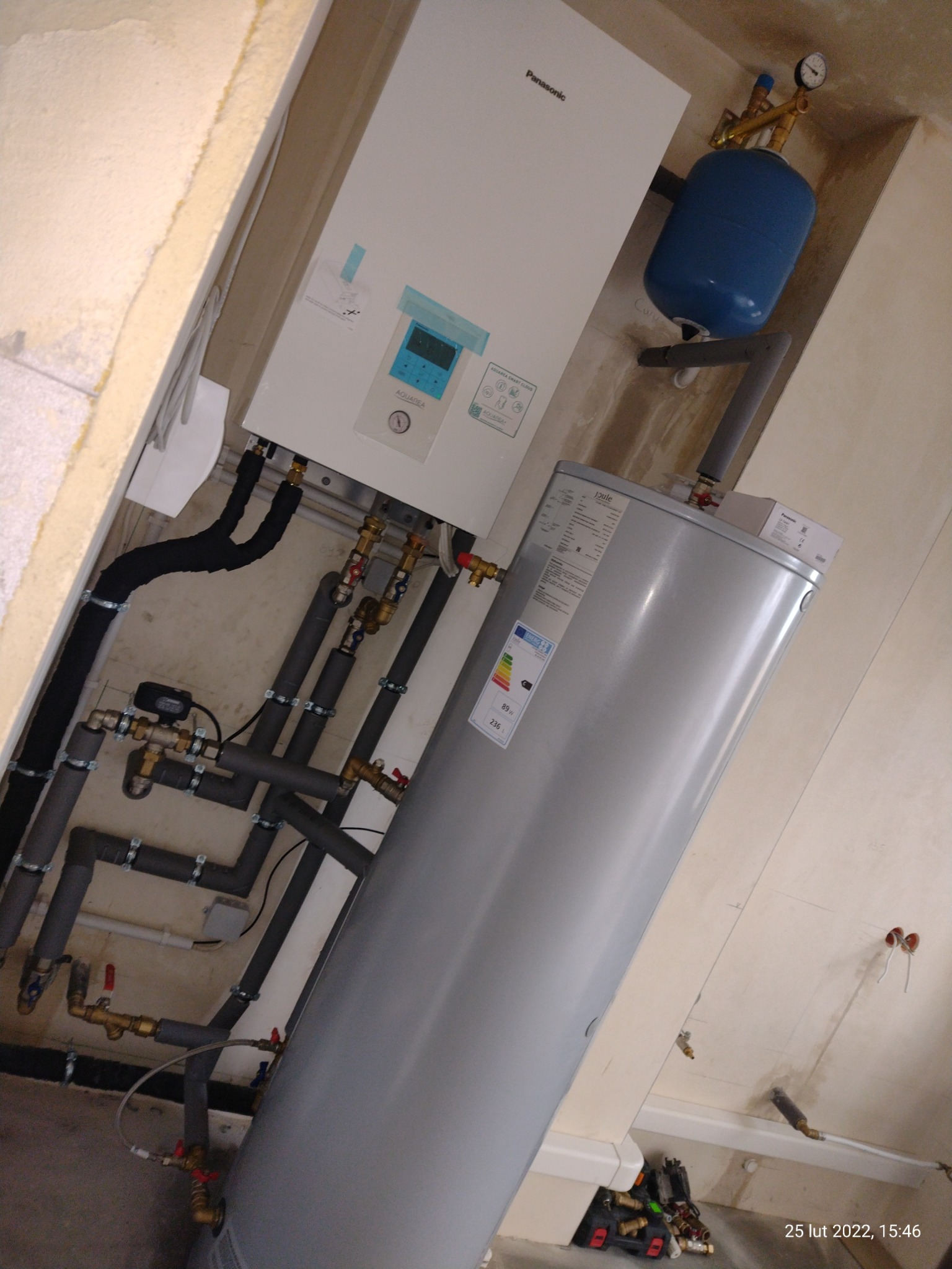 Instalacja kotła grzewczego Panasonic z zasobnikiem ciepłej wody i niebieskim zbiornikiem wyrównawczym, widoczne rury i zawory, na ścianie widoczne ślady po instalacji.