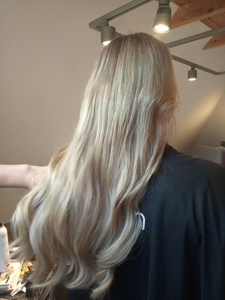 Długie, blond włosy z delikatnymi falami, widoczne od tyłu, oświetlone lampami sufitowymi. W tle fragment pomieszczenia z drewnianym stropem.