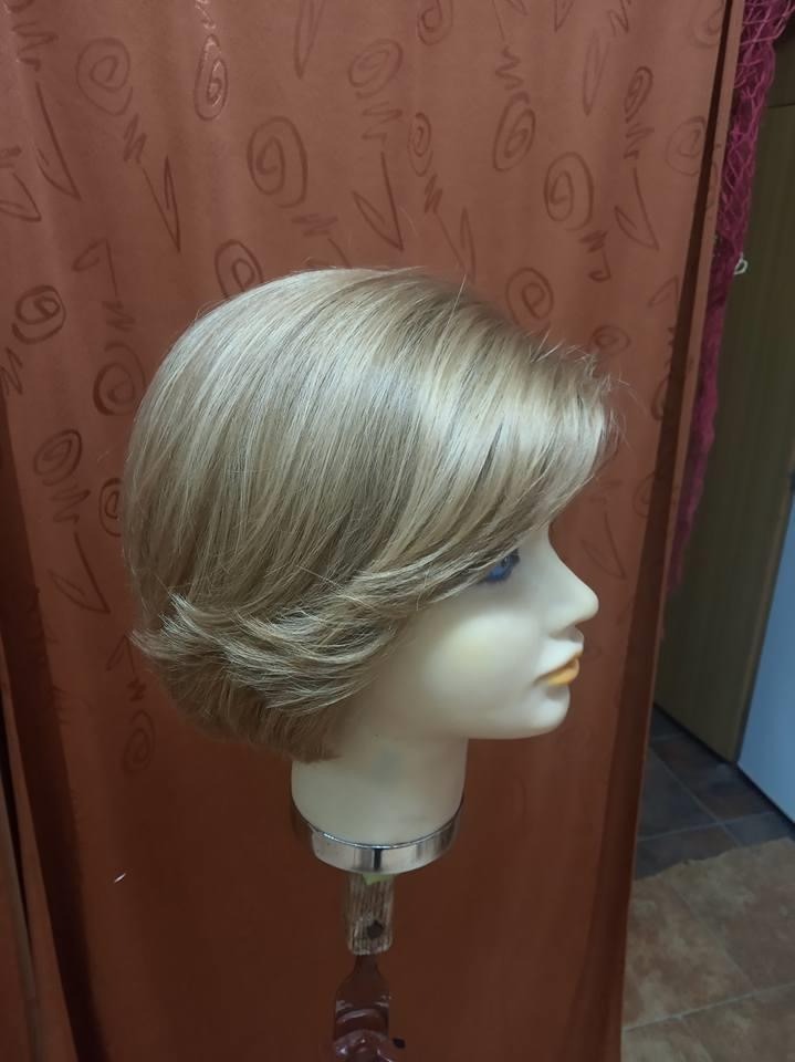 Krótka, blond peruka z grzywką na manekinie z tworzywa sztucznego, profil z widocznymi niebieskimi oczami manekina na tle pomarańczowej zasłony.