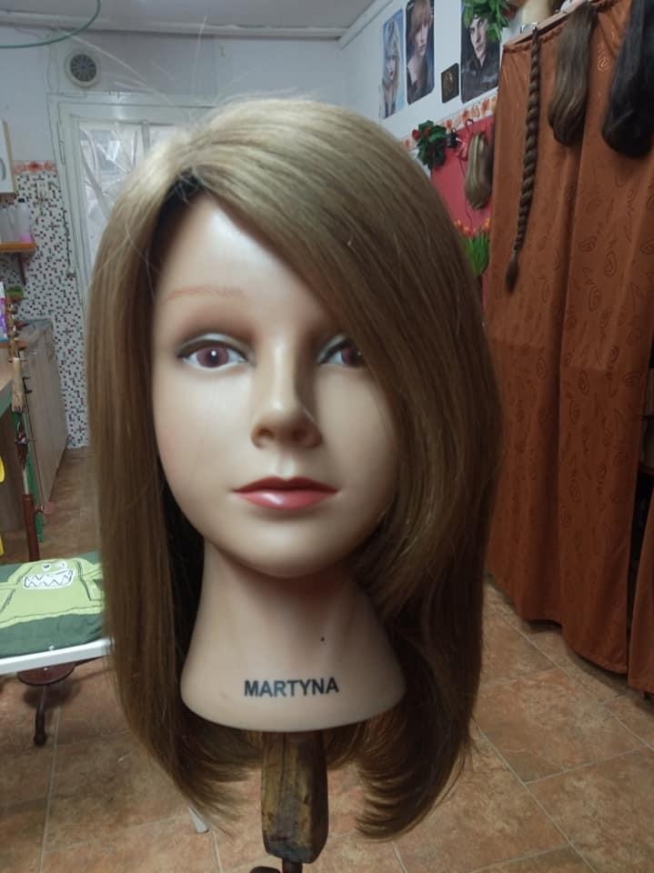 Manekin głowy z peruką z prostych, jasnobrązowych włosów do ramion, z grzywką na bok, na drewnianym stojaku, z napisem 'Martyna' na szyi manekina.