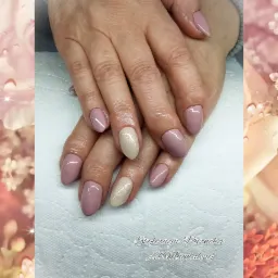 Dłonie z świeżo pomalowanymi paznokciami w odcieniach pudrowego różu i beżu z brokatem, ułożone na białym podkładzie, w tle rozmyte kwiaty.