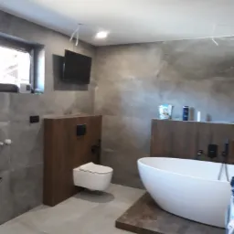 Nowoczesna łazienka z szarą, teksturowaną ścianą, podwieszaną toaletą i wolnostojącą białą wanną na drewnianym podeście; widoczne niedokończone podłączenia elektryczne.