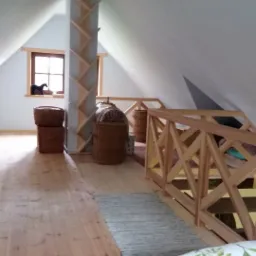 Poddasze z drewnianą podłogą, skośnymi ścianami, drewnianą barierką, regałem w kształcie drzewa, wiklinowymi koszami i łóżkiem z niebieską pościelą w kwiaty.