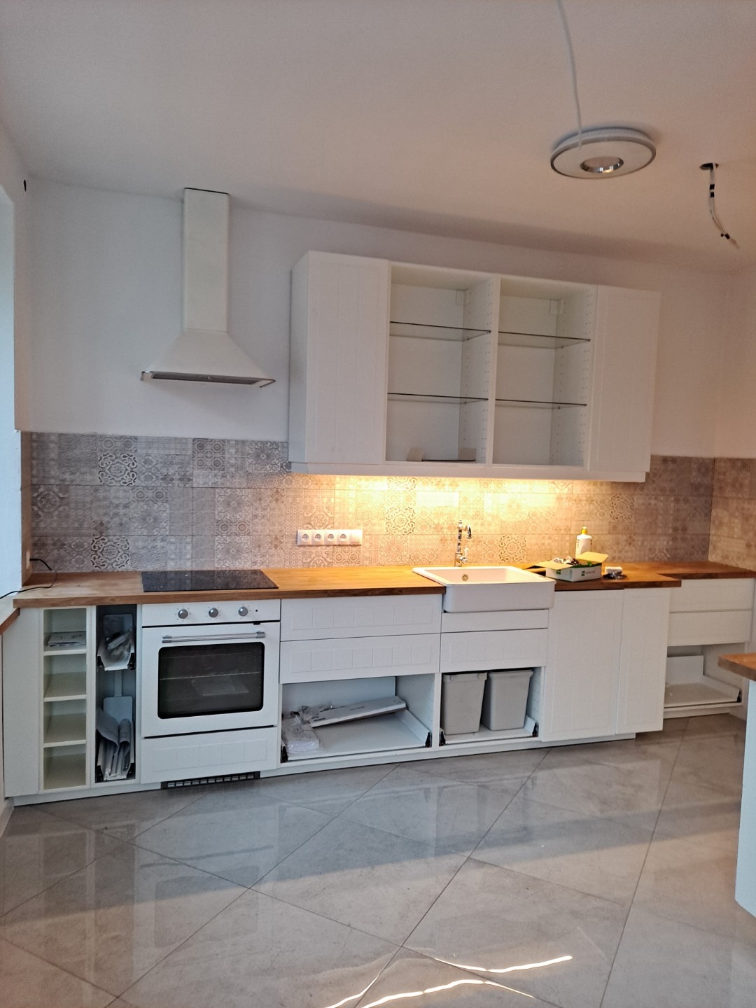 Biała zabudowa kuchenna z drewnianym blatem, widoczne otwarte szafki z akcesoriami, płytki dekoracyjne na ścianie i błyszcząca podłoga z płytek.