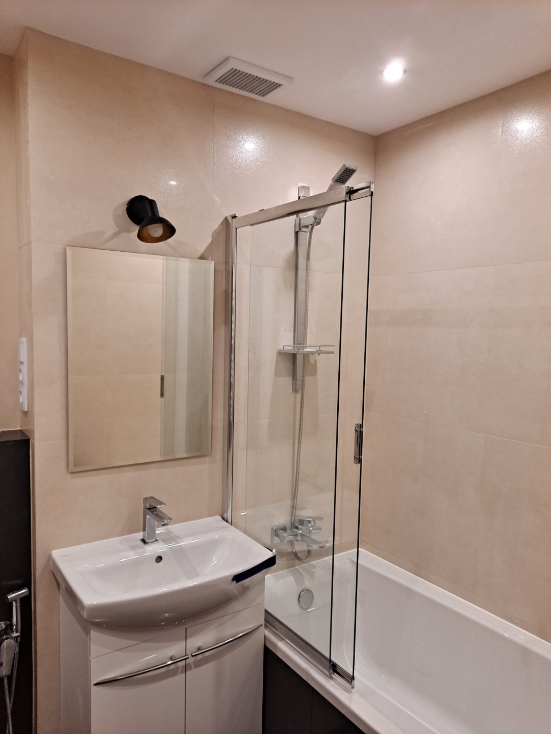 Jasna łazienka z beżowymi płytkami, kabiną prysznicową z przezroczystego szkła, białą umywalką z szafką i czarną lampką nad lustrem.