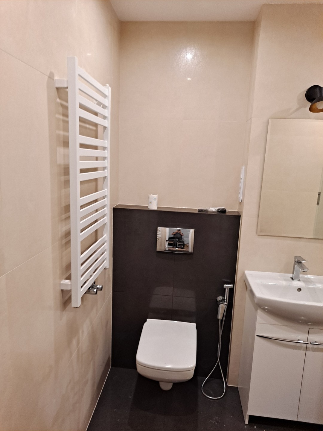 Nowoczesna łazienka z beżowymi płytkami, ciemnobrązową ścianą za toaletą, białą umywalką z szafką, grzejnikiem drabinkowym i bidetką.