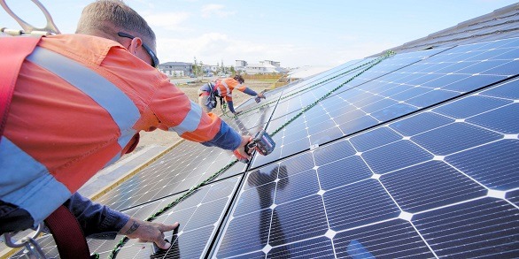 Montaż paneli słonecznych na dachu domu jednorodzinnego; pracownicy w odzieży roboczej zabezpieczeni linami, instalują kolejne panele, widoczne narzędzia pomiarowe i część zabudowy osiedla w tle.