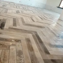 Ułożona pod kątem podłoga z płytek imitujących drewno w jasnych odcieniach szarości i beżu, widoczne ślady po fugowaniu, surowy stan ścian i widoczne przewody.