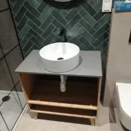 Nowoczesna łazienka z umywalką nablatową na drewnianej szafce z szarą płytą, zielone płytki ułożone w jodełkę na ścianie, widoczny fragment kabiny prysznicowej.