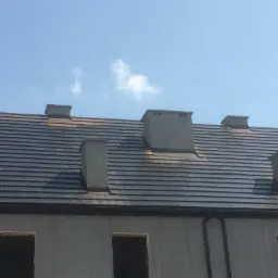 Nowy dach z ciemnej dachówki ceramicznej na budynku w trakcie budowy, widoczne cztery kominy, błękitne niebo w tle.