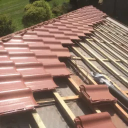 Montaż brązowej dachówki ceramicznej na drewnianej konstrukcji dachu, widoczne membrany dachowe i częściowo ułożone pokrycie z perspektywy z góry.