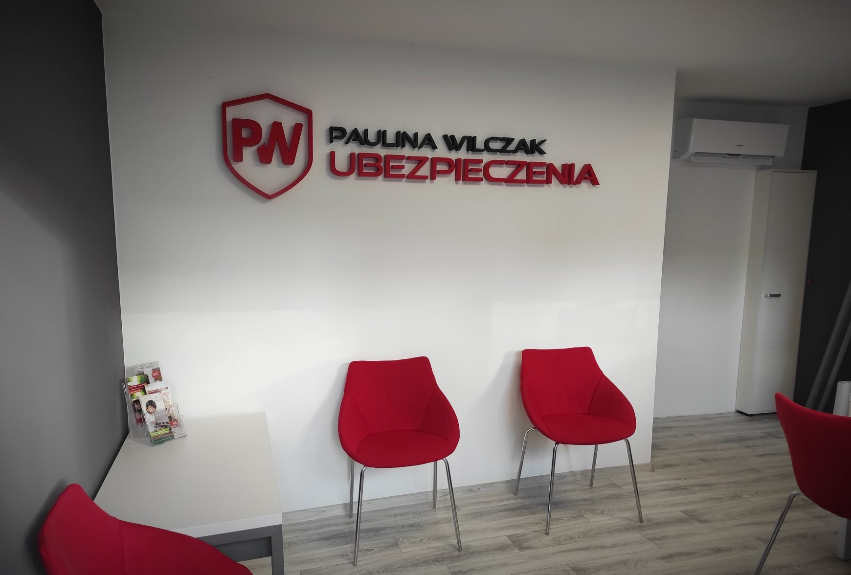 Wnętrze biura firmy ubezpieczeniowej Paulina Wilczak Ubezpieczenia w Lublinie z czerwonym logo i napisem na białej ścianie, trzema czerwonymi krzesłami i stolikiem z ulotkami.