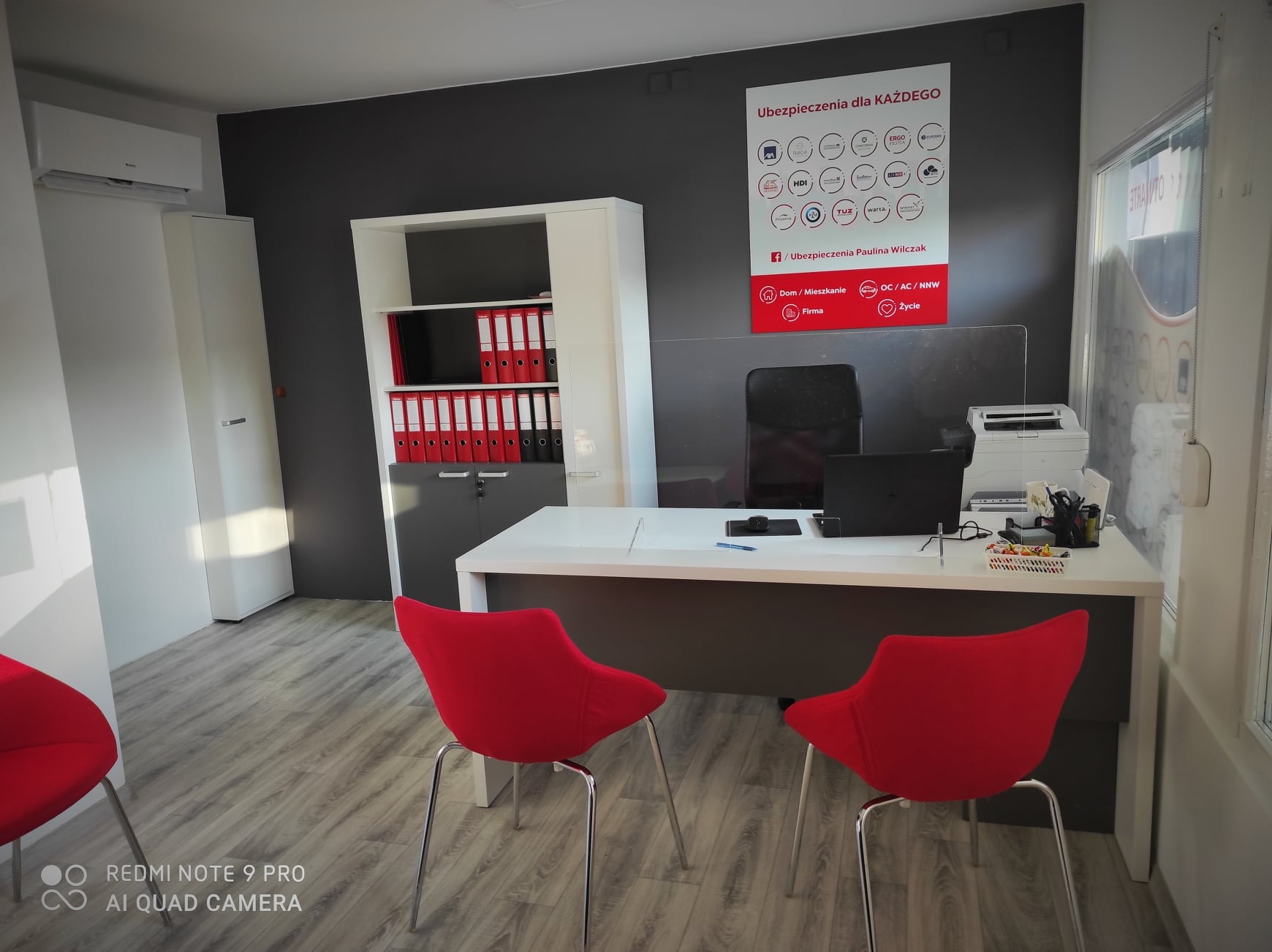 Biuro agenta ubezpieczeniowego z białym biurkiem, dwoma czerwonymi krzesłami i plakatem informacyjnym o różnych rodzajach ubezpieczeń.