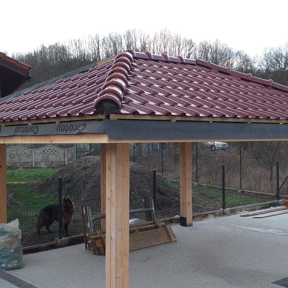 Zadaszenie z czerwoną dachówką ceramiczną w trakcie budowy, widoczne drewniane słupy konstrukcyjne, membrana dachowa z logo producenta oraz pies rasy owczarek niemiecki w tle.