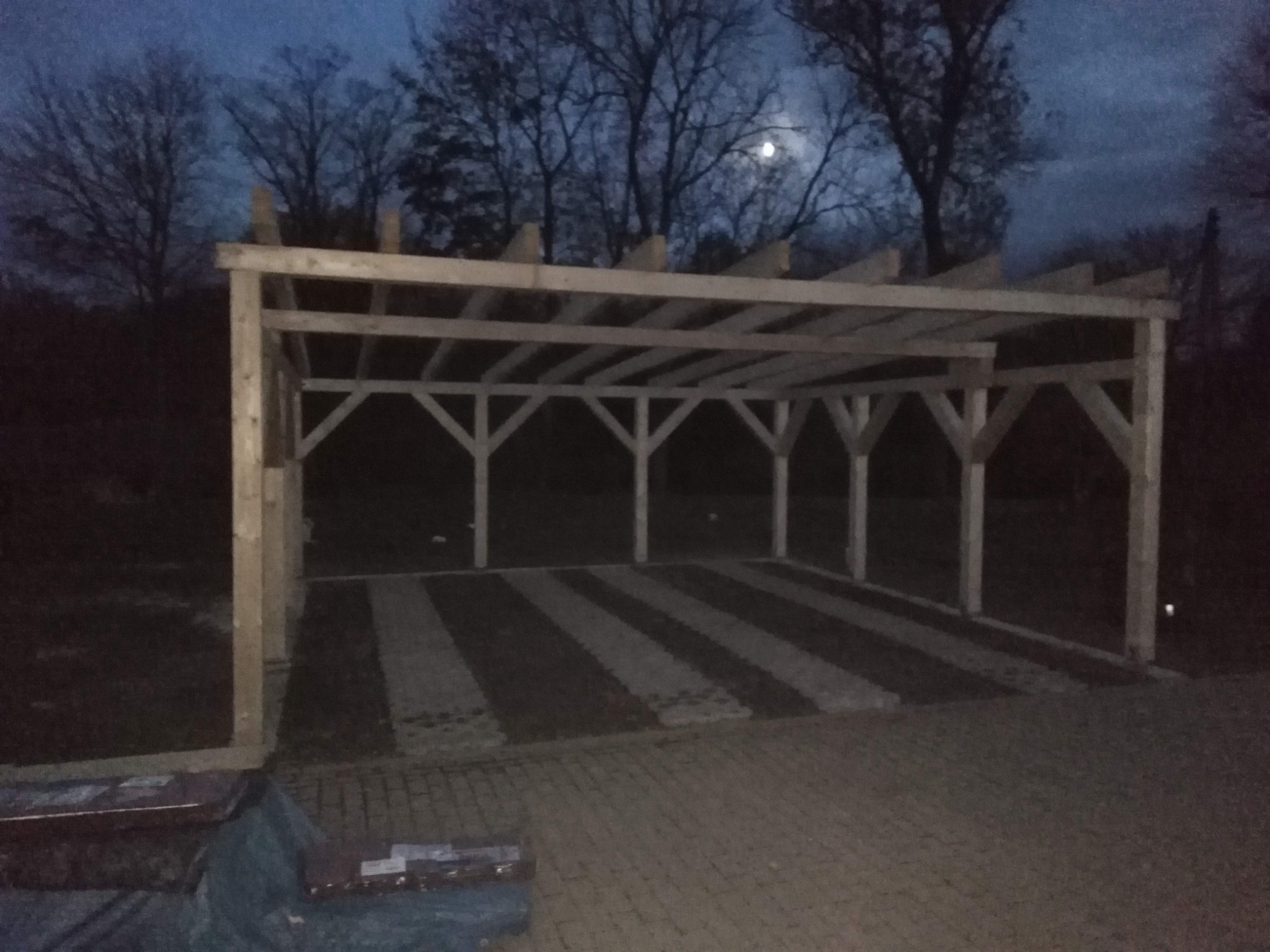 Surowa drewniana konstrukcja zadaszenia w trakcie budowy, widoczna w nocy na tle drzew i księżyca, z przygotowanym podłożem z kostki brukowej.