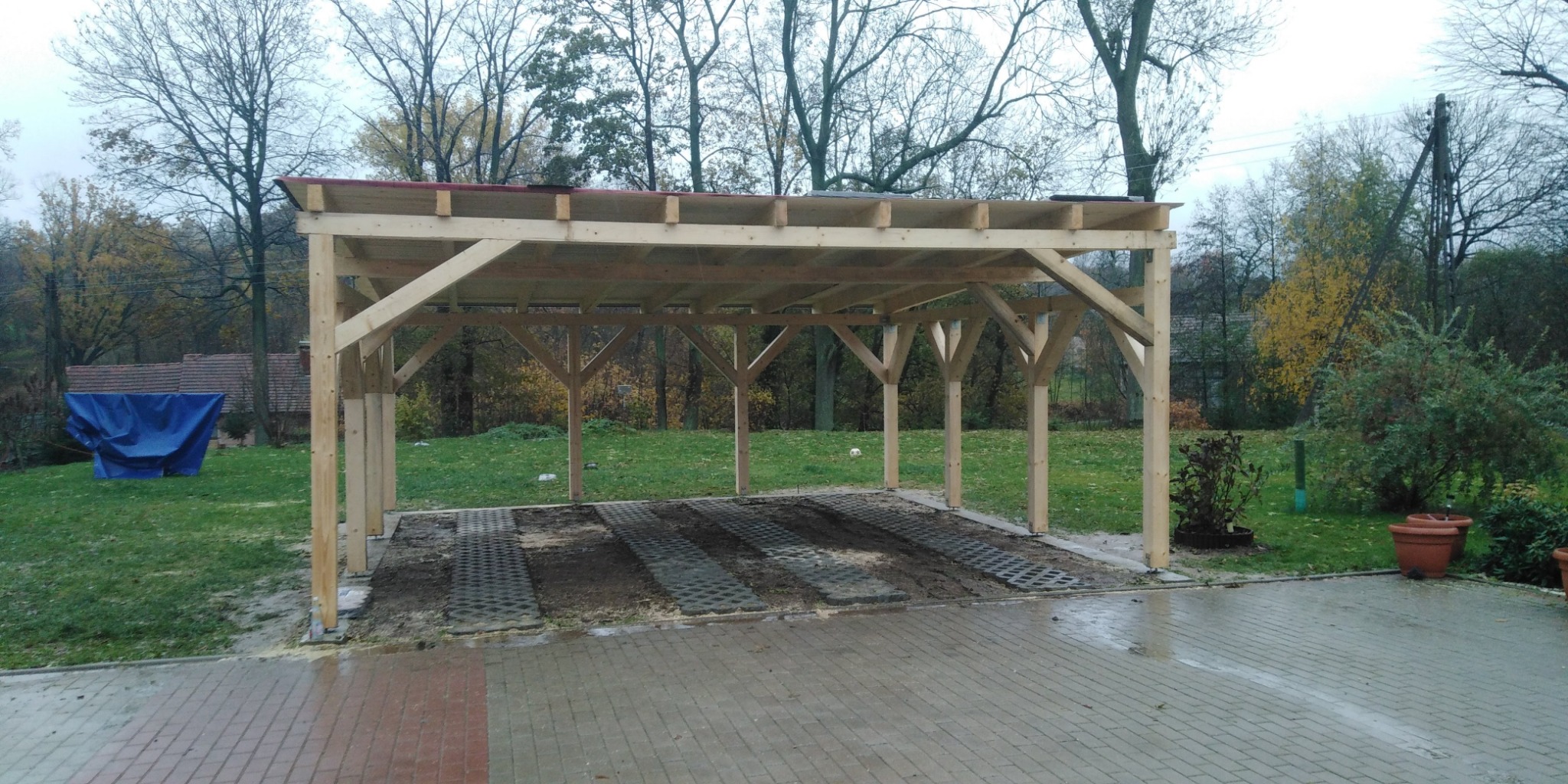 Surowa drewniana konstrukcja z częściowo ułożoną kostką ażurową na podjeździe, widok na ogród i dom w tle.