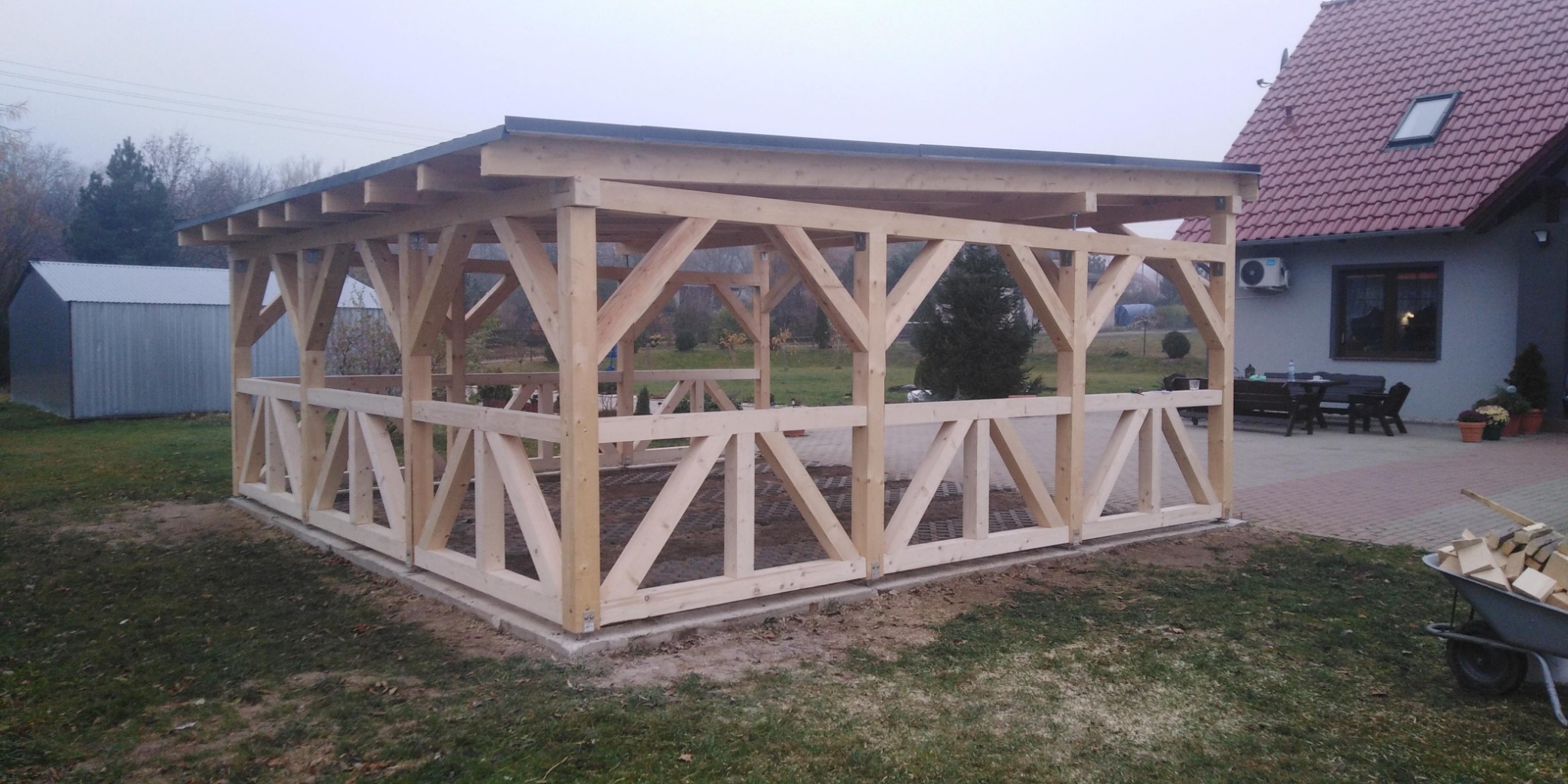 Drewniana konstrukcja altany w trakcie budowy, widoczne elementy dachu i ozdobne kratownice ścian, w tle dom z czerwonym dachem i wózek z drewnem.