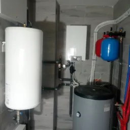 Wnętrze kotłowni z zamontowanym białym zasobnikiem ciepłej wody, szarym bojlerem, wiszącym piecem gazowym i dwoma zbiornikami wyrównawczymi w kolorach czerwonym i niebieskim, całość na tle szarych...