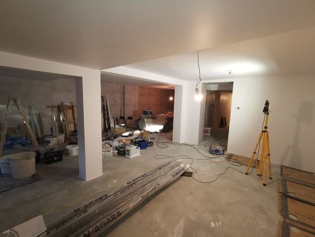 Wnętrze w trakcie malowania: widoczne świeżo pomalowane ściany w kolorze białym, liczne narzędzia malarskie, drabina, lampa na statywie i materiały budowlane na podłodze. Częściowo pomalowane...