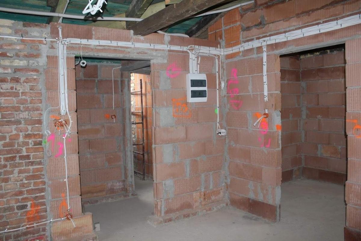Instalacja elektryczna w surowym wnętrzu, widoczne kable prowadzone po ścianach z cegły, skrzynka elektryczna i oznaczenia w jaskrawych kolorach.