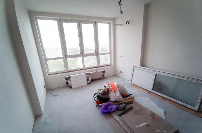 Pokój w stanie deweloperskim przed malowaniem; widoczne przygotowane ściany, okno z widokiem na miasto, grzejniki, materiały budowlane i puszki elektryczne na ścianie.