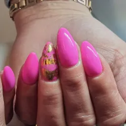 Ręka z jaskrawo różowym manicure hybrydowym, jeden paznokieć z efektem złotej folii, w tle złota bransoletka.