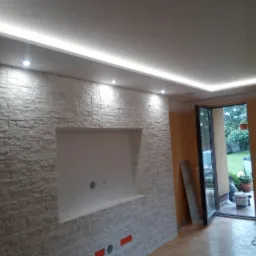 Wnętrze pokoju z dekoracyjną ścianą z imitacji kamienia, wnęką na telewizor i oświetleniem LED podsufitowym, widok na ogród przez otwarte drzwi balkonowe.