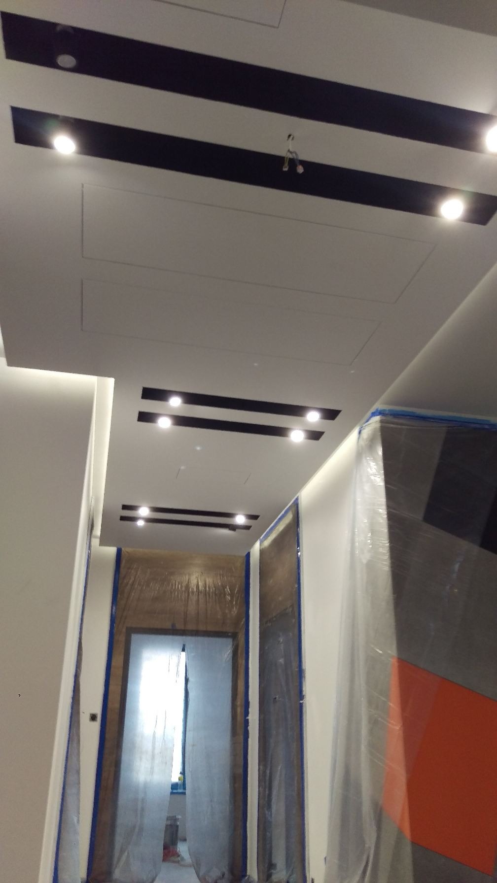 Przygotowania do prac wykończeniowych w mieszkaniu: Sufit z czarnymi wstawkami i punktowym oświetleniem, zabezpieczone folią drzwi i fragment ściany z pomalowanym wzorem.