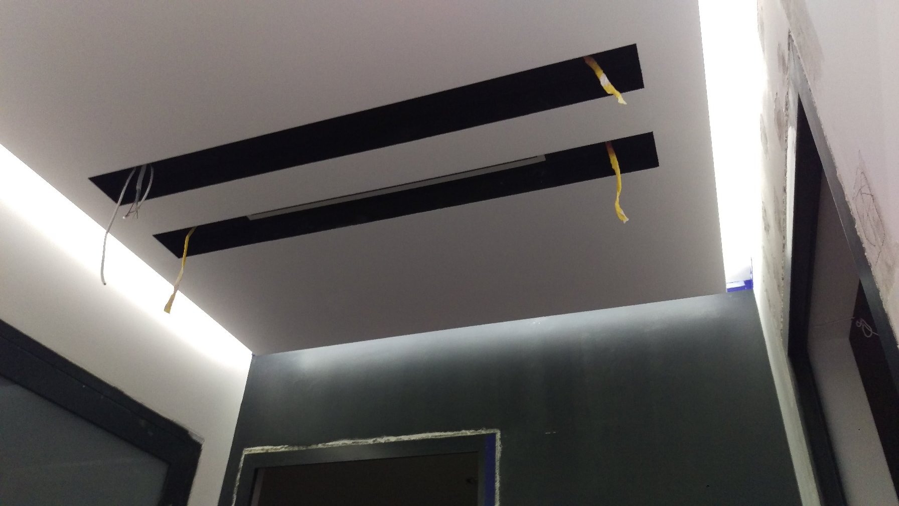 Podwieszany sufit z czarnymi wnękami i wystającymi kablami, oświetlenie LED wzdłuż krawędzi, szara ściana w tle.