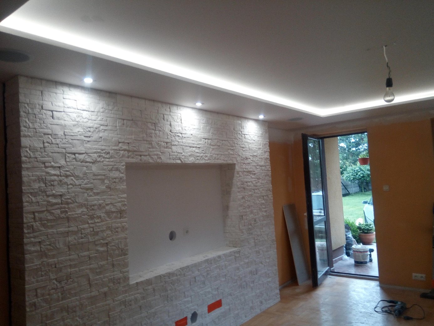 Wnętrze pokoju z dekoracyjną ścianą z imitacji kamienia, wnęką na telewizor i oświetleniem LED podsufitowym, widok na ogród przez otwarte drzwi balkonowe.