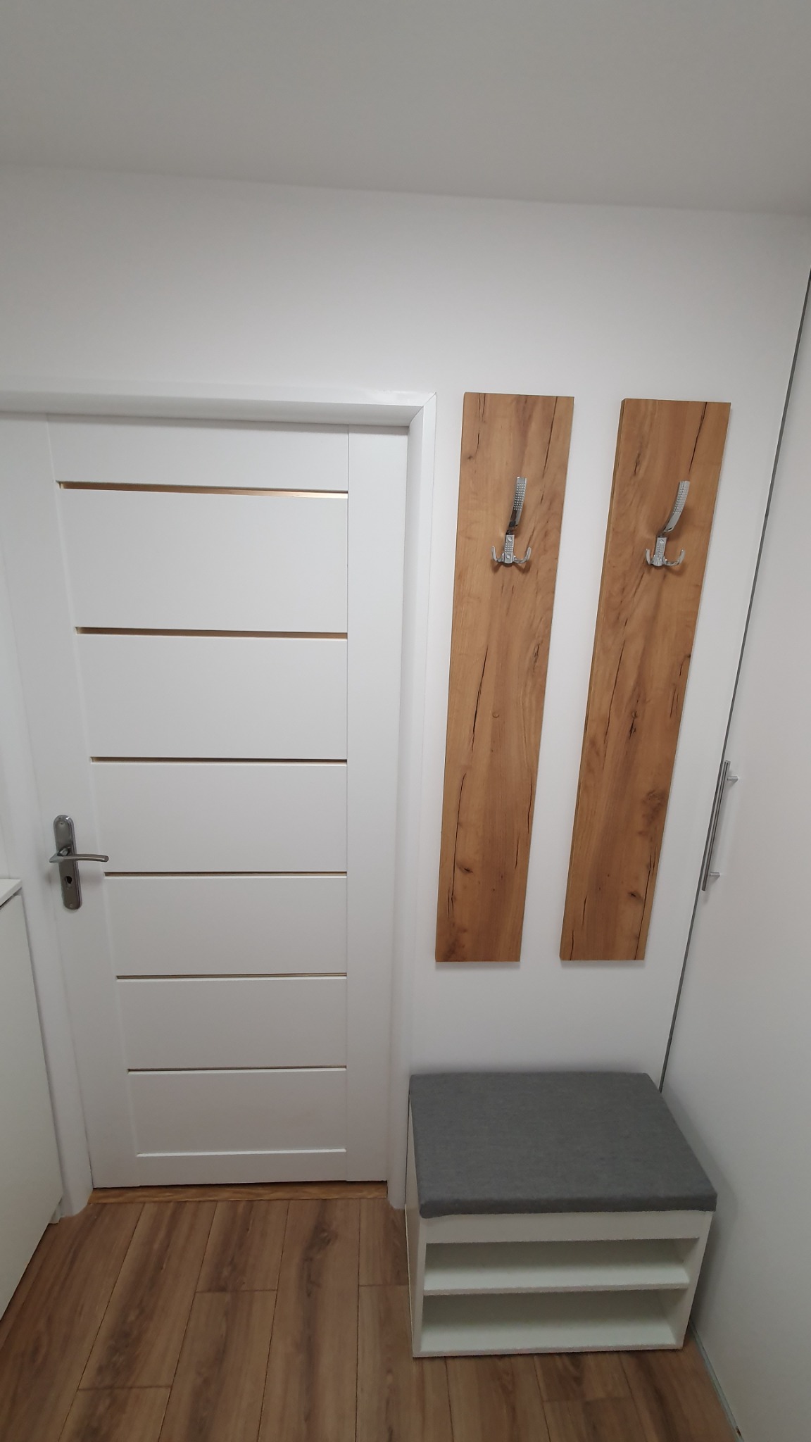 Białe drzwi wewnętrzne z poziomymi wgłębieniami, drewnopodobne panele ścienne z metalowymi wieszakami oraz biała szafka z szarym siedziskiem w przedpokoju.