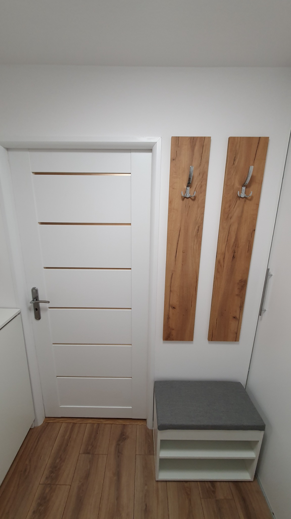 Nowoczesny przedpokój z białą, panelową drzwią z złotymi wstawkami, dwoma drewnianymi panelami z metalowymi wieszakami i białą szafką na buty z szarym siedziskiem.